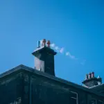 Expert Tips for Maintaining a Healthy Chimney System - Your Source for the Latest Housing Trends | Housing Market News| A plume of smoke rises from a chimney system on the roof of a stone building under a clear blue sky.